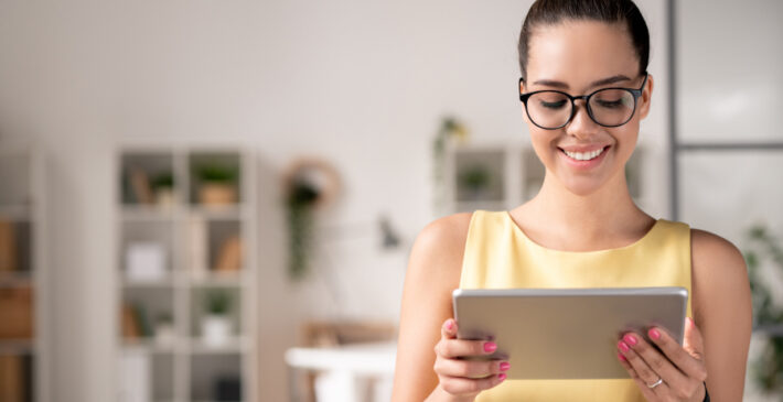 Smiling young project manager in eyeglasses using digital tablet while working with online platforms