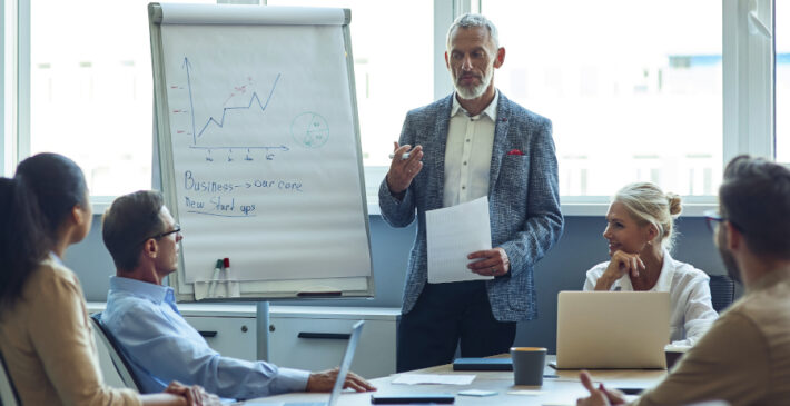 Confident mature businessman giving a presentation, explaining something to colleagues while having a meeting in the office. Business people working together